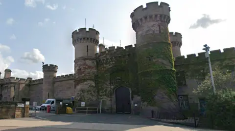 The entrance of HMP Leicester, in Welford Road, Leicester. It is a stone-built prison with walls with castle-like designs at the top including circular towers with arrow slits in them. Ivy is growing across the front of the prison walls.