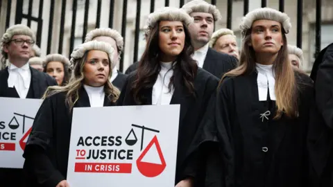 A wide shot of a group of barristers wearing black robes, white collars and barrister wigs stand in front of a black iron gate outside of Laganside Courts in Belfast. One is holding a white sign with 'Access to Justice in Crisis' written in black and red text.