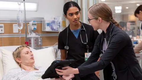 HBO Max Unknown actor lying in hospital bed with Dr Samira (Supriya Ganesh, centre) and Dr Mel (Taylor Dearden, right) at her bedside showing her a screen on a tablet