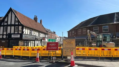 Richard Madden/BBC Yellow barriers and traffic cone lining the street, with a large, red "ROAD CLOSED" sign in front. Shops and businesses can be seen behind the barriers. The sky is blue.
