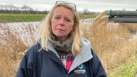 Bryony Sadler. She has long blonde hair and is pictured outside in Somerset. She is wearing a navy blue coat with a grey fluffy lining and a green scarf. She has glasses on her head. Behind her are pumps, pumping flood water out.