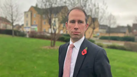 Thames Valley Police and Crime Commissioner Matthew Barber looks at the camera. He is standing in a green area on the housing estate Great Western Park - there are trees and buildings in the background. He is wearing a grey suit, a pink-coloured tie with dots and there is a poppy pinned on his lapel.