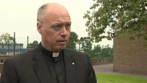 El padre Martin Graham está de pie junto a un edificio de ladrillo, con un árbol a la derecha. Lleva su collar de perro y su hábito sacerdotal negro. Tiene el pelo ralo y ojos azules.