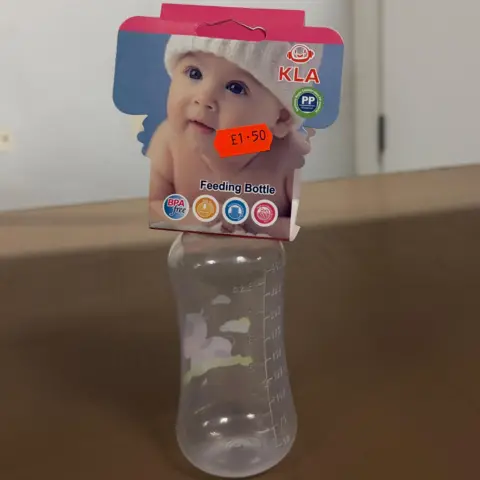 Jasmine Lowe/BBC News A plastic baby bottle placed on a shelf. The bottle has been deemed dangerous by trading standards. It does look cheap and slightly tacky and there is a sticker with the price £1.50 on the packaging. The packaging is blue and it has a large picture of a new-born baby in a woollen hat. It says feeding bottle on the top. 