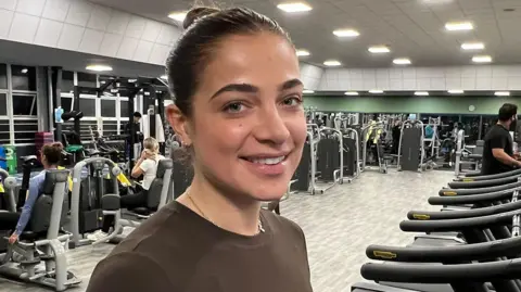 Vikki has dark brown hair tied up at the back of her head in a bun. She is wearing a brown long-sleeved exercise top and is standing on a treadmill in a spacious gym, which is filled with cardio and strength equipment. Rows of treadmills are lined up on the right, while several people use weight machines in the background on the left. The gym has bright overhead lighting, grey flooring, and large windows along one wall.