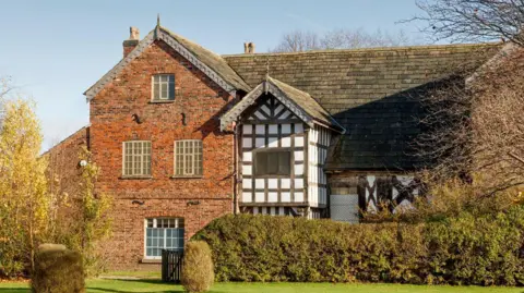 A medieval-style building can been seen, made both of red brick and traditional black and white Tudor architecture. A neat lawn and bushes are visible also 