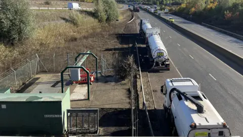 Tony Fisher/BBC Three tankers can be seen parked at the side of the A421 next to a pumping station. They have long hoses that are stretched to the pumping station. Cars can be seen in the distance on the carriageway.