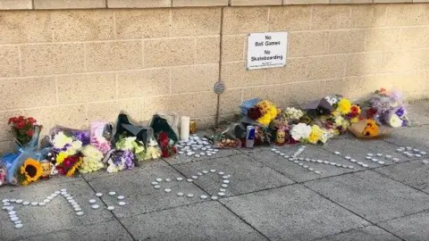 Jon Wright/BBC Bunches of flowers and candles are placed against a wall. Cans of drink are left too. Tealights spell out the names Mo and King with a heart depicted by tealights.