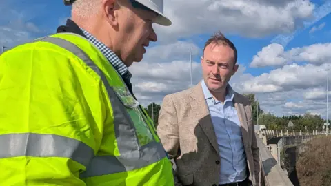 Blake Stephenson Blake Stephenson and another man standing outside on what appears to be a road overpass. Stephenson is wearing a light coloured blazer and blue shirt. Another unidentified man is wearing a high visibility jacket and a white hard hat. The collar of this man's blue checked shirt is just visible. The two men appear to be in conversation with one another. The road can be seen disappearing off into the background. It is a bright, sunny moment, though there are puffy white clouds in the sky.