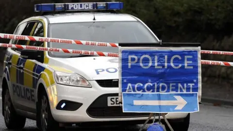 A police car is parked behind two red 'do not cross' police tape lines and a blue 'accident sign'