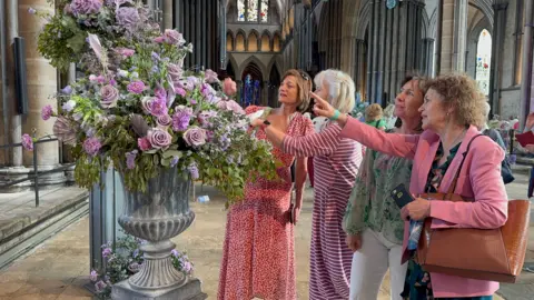 A arrangement of pink and purple flowers in a stone vase in the middle of the cathedral's main hall. Four women wearing pink and red clothes are looking and pointing at the flowers