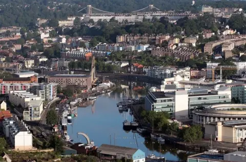 An aerial view of the centre of Bristol with the floating harbour in the foreground and the Clifton Suspension Bridge in the distance