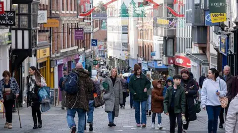 Getty Images Shoppers including men, women and children are walking in a pedestrianised street in Shrewsbury with shops on either side. They are wearing coats and hats in the autumn and are talking and looking around them.