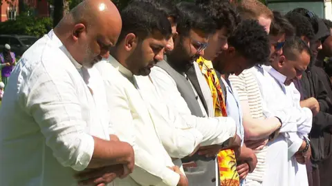Men in white kaftans standing in a line with heads bowed and arms folded across their chests. They are standing in a grassy field on a sunny day.