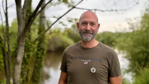 A man with a balding head and grey beard wears a khaki T-shirt and stands in front of a river and some trees.