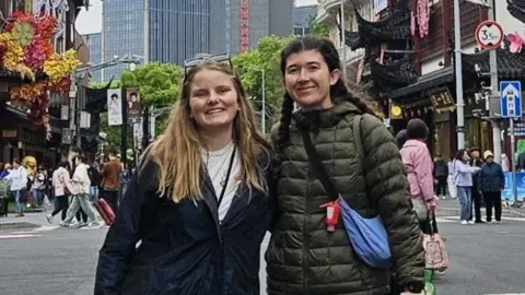 The Jo Cox Way Two women in their 20s with their arm around each other smile at the camera. There is a cityscape behind them, possible japan 