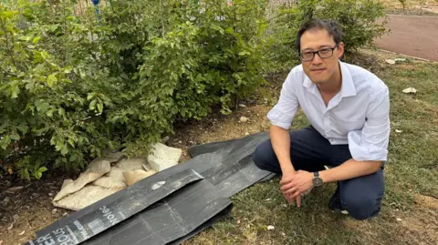 Johnny Luk wearing a white shirt and dark trousers crouches beside a small pile of discarded black plastic panels and rubble on a grassy verge next to shrubs. A paved path runs behind the bushes in an outdoor landscaped area.