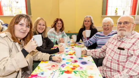 Six people, five women and one man, are sitting around a table, holding white mugs. Councillor Kelly Middleton is on the left, followed by a blonde woman, a woman with red hair, and two women with grey hair. The man has white hair and facial hair. 