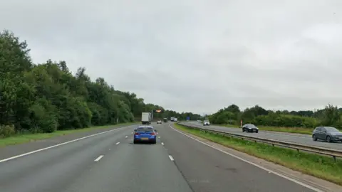 The M6 has six lanes in total, separated by a grassy central reservation with a low metal barrier running along it. The road is bordered by tall trees on the left, and smaller trees and shrubs on the right. There are a mix of cars, vans and trucks on the road, but they are fairly well-spaced apart.