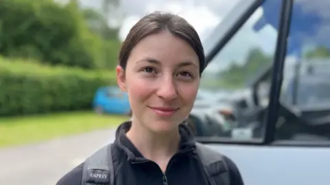 Daisy Drew, stood next to her camper van and smiling at the camera. She has black hair, tied in a pony tail, and is wearing a black fleece top, with a rucksack on her back.