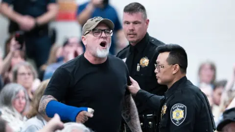 A man shouts as members of law enforcement escort him out of the building during a town hall meeting.