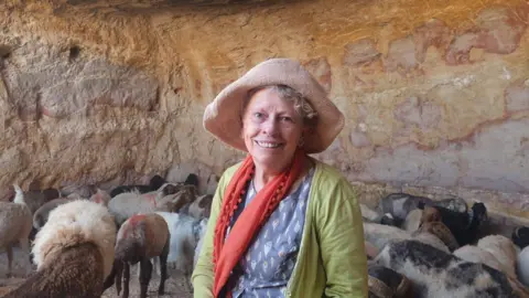 A woman smiling at the camera wearing a hat, red scarf, green jumper and a blue top. You can see a group of sheep behind her and she appears to be in a .cave