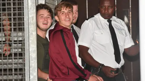 PA Media Albert Bowers and Jessie Cole smile as they are led in handcuffs by two prison officers