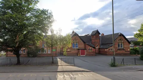 Google the front of Holmer Academy. it is a red brick school building with a grey roof and large concrete area for cars and a black metal fence. There is a road outside the fence