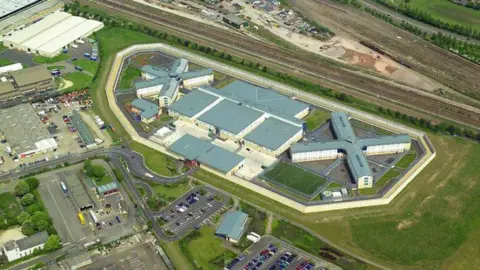 Alamy An aerial image of HMP Peterborough. The prison has several large buildings, all with a grey roof. It is immediately surrounded by a field with a railway line and industrial estate also nearby.