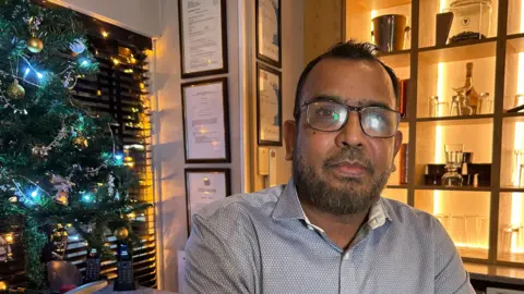 Mr Abu Taher Khan is a man of Asian descent who is wearing glasses. He has short black hair and a short black and grey beard. He is wearing a button down shirt. There is a Christmas tree to the left with lights on it. It is a head and shoulders shot.