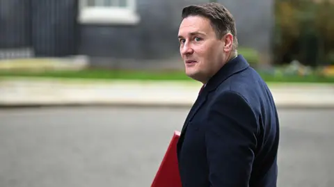 Getty Images A man in a dark navy coat looks back over his shoulder while walking, carrying a red folder under his arm. He is outdoors on a paved surface with a dark building entrance and greenery visible in the blurred background. His hair is short and dark brown, swept to one side. The image is taken from behind at a slight angle.
