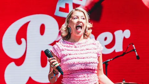 Getty Images Caity Baser performing on stage. She wears a frilly pink top, hoop earrings and red lipstick, her mouth open wide as she seems to scream in excitement towards the audience. The staging behind her is red, with 'L' plates and her name in a white font. 