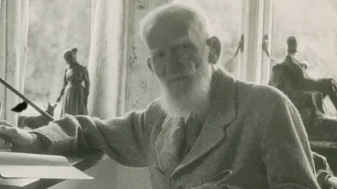 National Trust A grainy old black-and-white photo of George Bernard Shaw. He is dressed smartly and has a long white beard. He is sitting at a writing desk holding a pen.