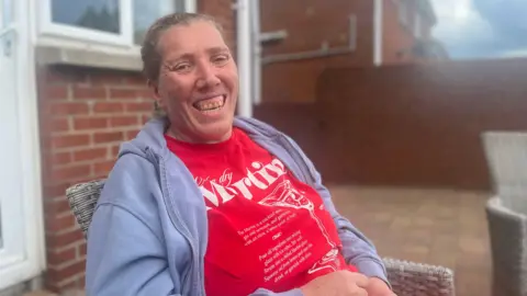 Laura is picture looking toward the camera and smiling. She's wearing a bright red t-shirt with white writing on it and a grey zip-up hoodie. She's sitting on a wicker chair outside a red brick home. 