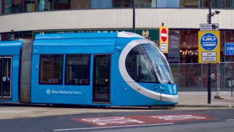 A blue West Midlands Metro tram on a street. The tram with West Midlands Metro branding is at a road junction, with partly yellow traffic signs and the pavement behind it.