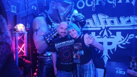 Lena Joyce A bald man and a woman with blue hair are standing in a bar with dim, blue lighting. There is a mural on the wall that reads "rock bar" and depicts a muscled man singing into a microphone with leather clothing. The woman is holding up a rock hand sign