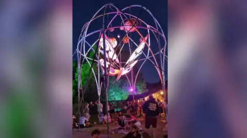 Keynsham Music Festival A huge metal butterfly is suspended from a dome created from hoops of metal. Planet-like spheres hang above the butterfly. It is night time and a purple light is shining on the artwork. 