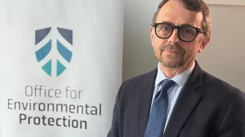 Robbie McDonald, wearing a dark suit jacket, blue shirt, dark blue tie and black-rimmed glasses, looks towards the camera.   He is standing in front of a branded sign which says "Office for Environmental Protection."