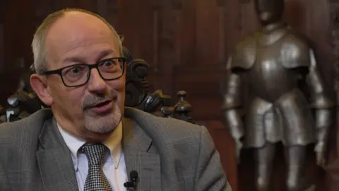 A man with glasses and a beard sits on an ornate chair in a wood panelled room with a metal suit of armour behind him