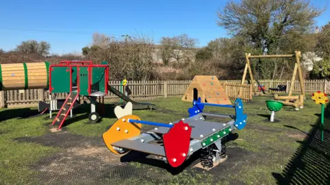 Joanna Taylor/BBC Weston Colville playground on a sunny day. In the foreground is a large seesaw. Behind it is the side of the Lancaster Bomber climbing frame, a small wendy house with climbing pads on the side and a swing set. There is patchy grass beneath and trees in the background. 