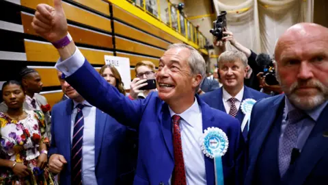 Clodagh Kilcoyne/Reuters A group of people at an election count. In the middle is Nigel Farage with his thumb in the air.