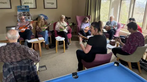 Ellie Colton/BBC A group of eight people sat in a circle on chairs playing the ukelele