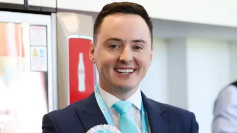 Darren Grimes is smiling at the camera. He is wearing a navy suit with a light blue tie and a Reform rosette in the same colour. His short brown hair is swept to the side. 