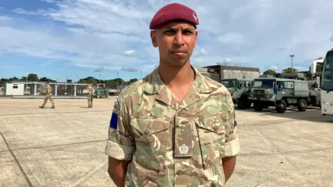Henry Godfrey-Evans/BBC A serious-looking man in a camo uniform and a red beret with his hands behind his back.