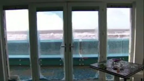 The glass of double doors and windows has been blown out. Glass is scattered across the floor. Beyond the doorframe, you can see a concrete wall and the waves of the sea further in the distance. 