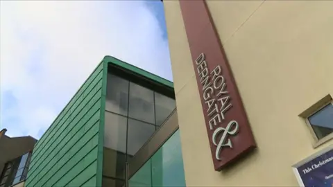 The outside of the theatre with a vertical maroon sign saying "Royal and Derngate" going up the side of the building. The shot has been taken from a low angle, looking up towards a cloudy sky. 