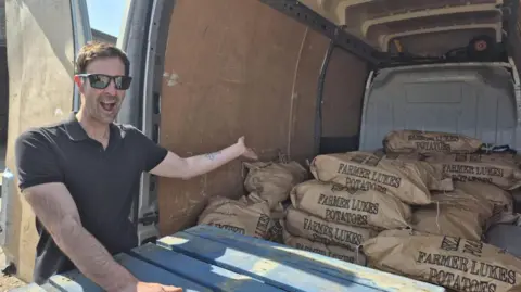 Luke Abblitt Luke, a man, with short brown hair. He is wearing a black T-shirt and black sunglasses and is gesturing towards brown sacks of potatoes in the back of a van. The bags all say "FARMER LUKES POTATOES".
