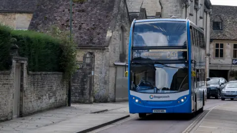 A blue bus with 'Cirencester' as the destination on it drives through a single laned road next to a quaint looking brick wall.