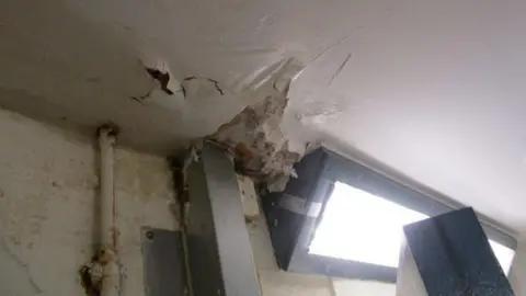 HM Inspectorate of Prisons White ceiling with peeling paint and visible water damage, some material falling down out of the roof.