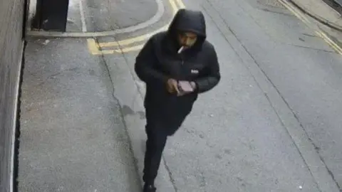 GMP Rehman, dressed in black with his hood up, walking along a pavement - as caught on CCTV.